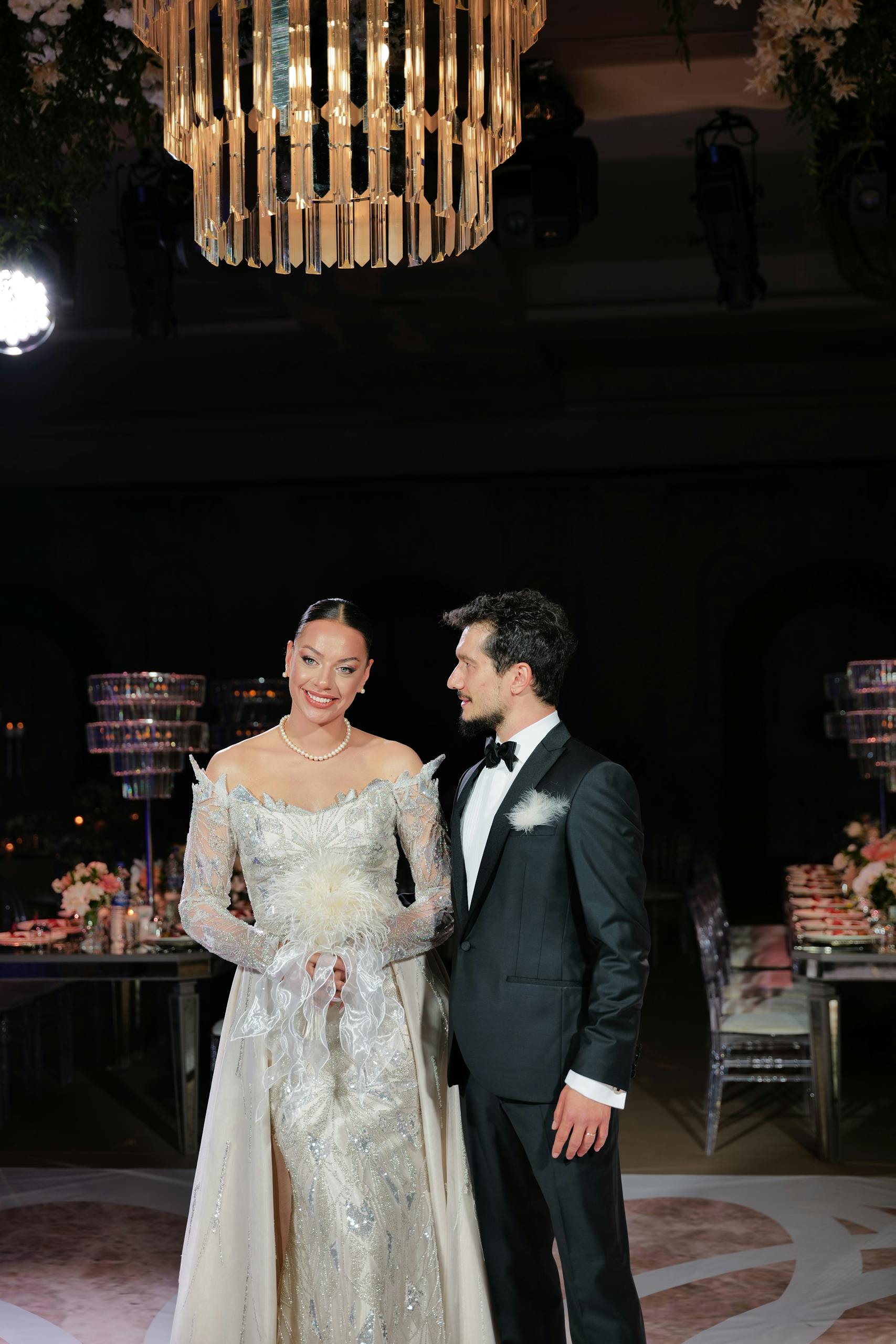 Bride and groom posing elegantly under a glamorous chandelier during their wedding.
